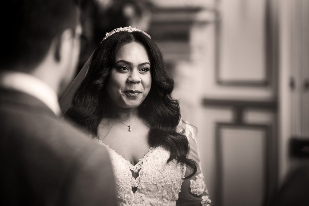 A bride in a lace wedding dress and veil looks at a person in a suit, smiling during a wedding ceremony indoors. Wedding Photo taken by Jennings Photography