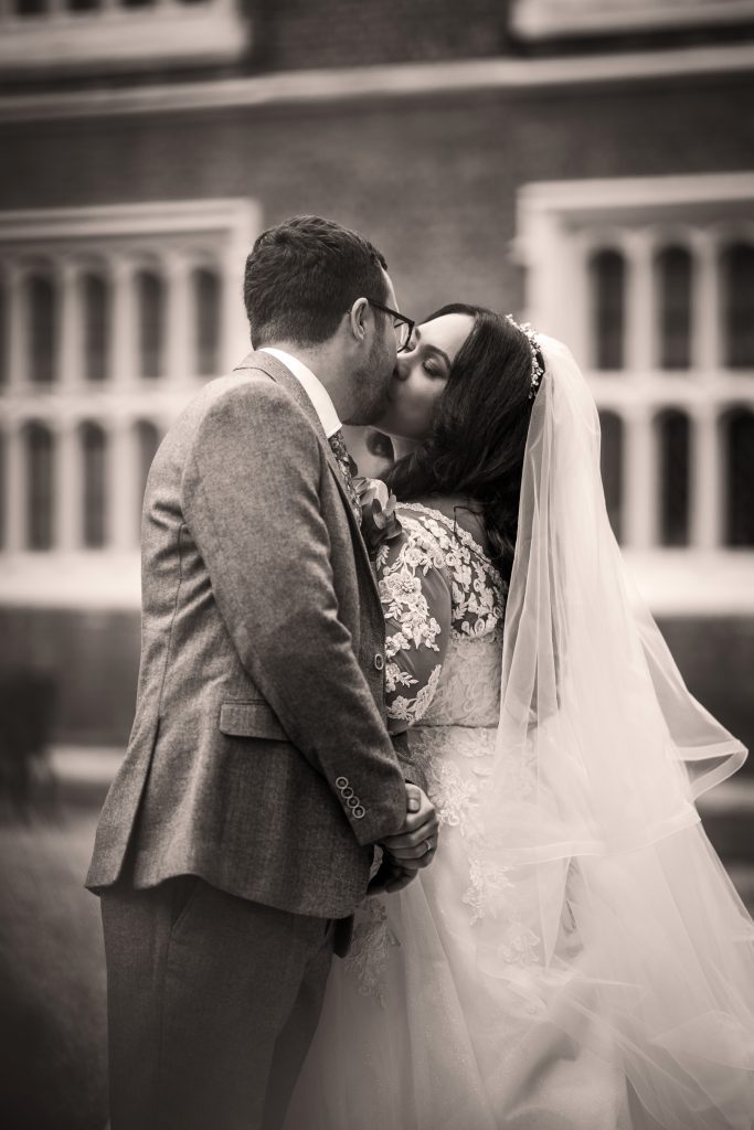 A bride and groom kiss outdoors in front of a building with large windows. The bride wears a lace gown and veil; the groom is in a suit. Wedding Photo taken by Jennings Photography