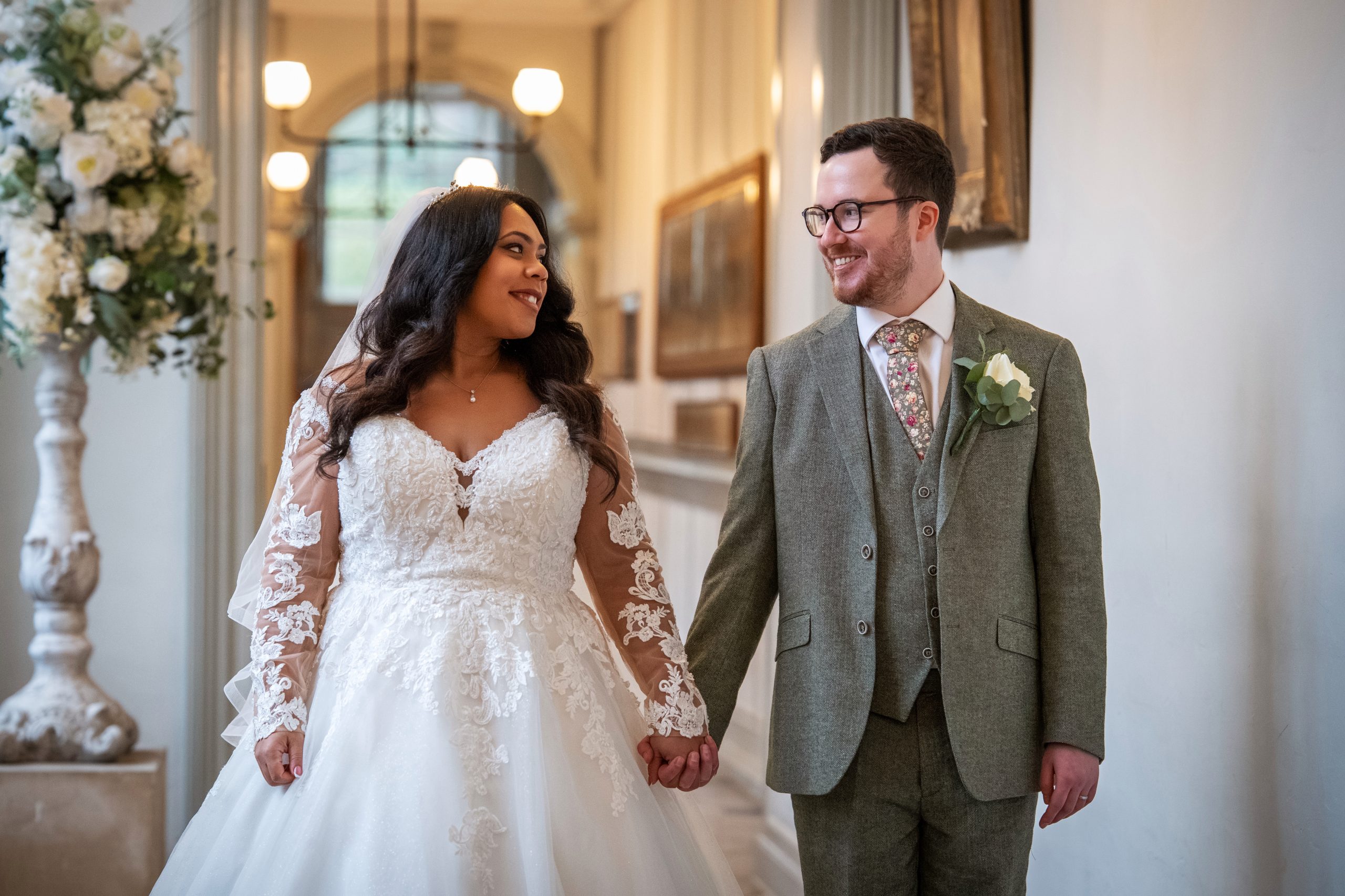A bride in a white lace wedding dress and a groom in a gray suit holding hands and smiling at each other while walking indoors. Wedding Photo taken by Jennings Photography