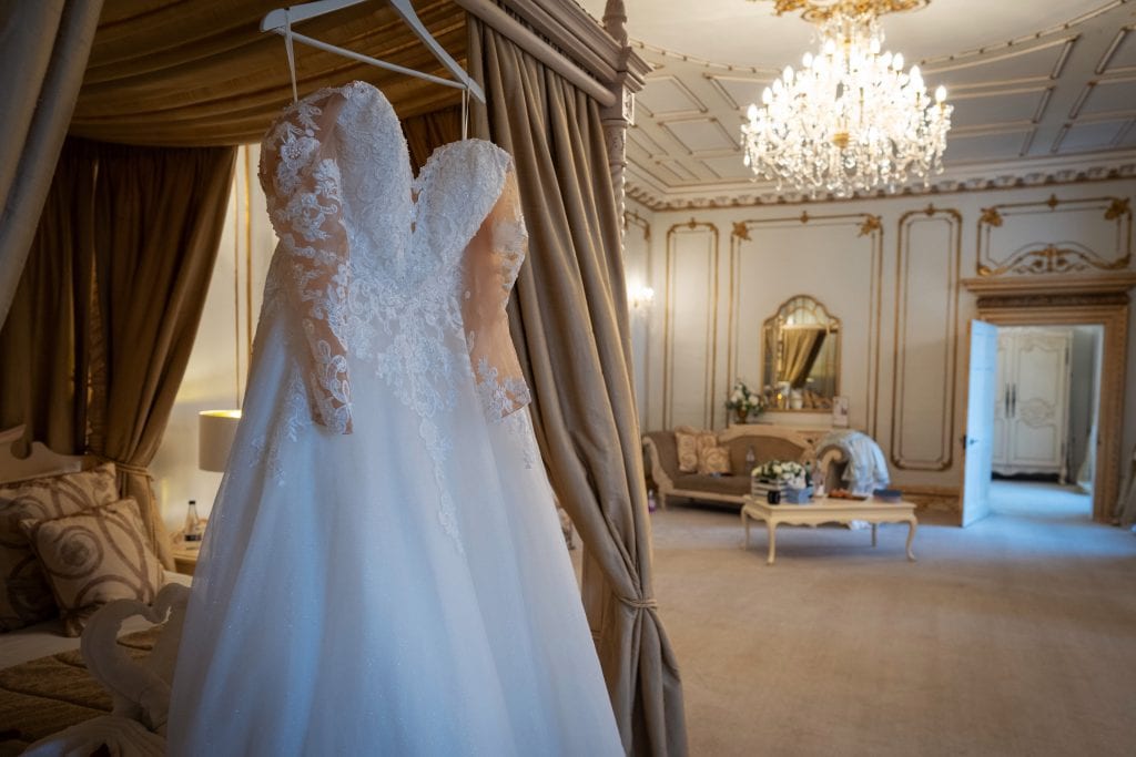 A white lace wedding dress with sheer sleeves hangs on a canopy bed in an elegant, ornate room featuring chandeliers and antique-style furnishings. Wedding Photo taken by Jennings Photography