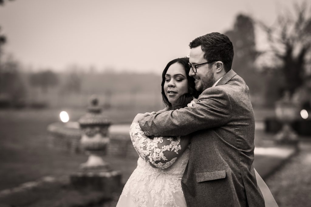 A couple in wedding attire embraces outdoors on a cloudy day, with blurred trees and stone decorations in the background. Wedding Photo taken by Jennings Photography