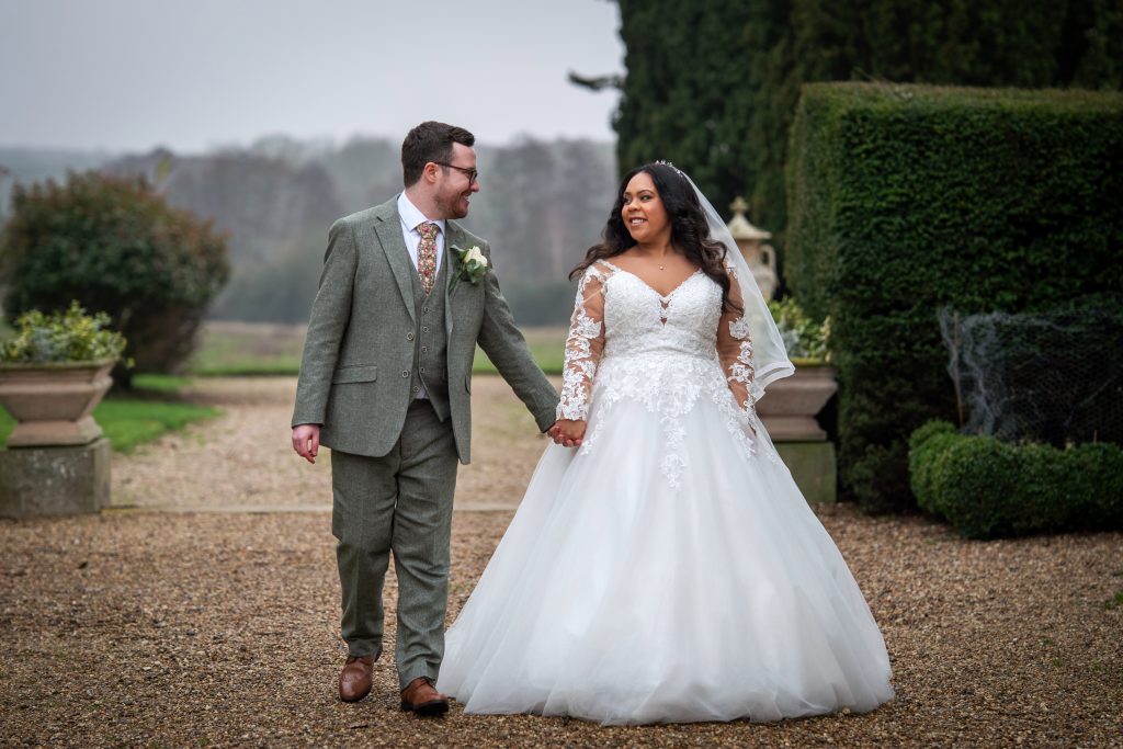 A bride in a white gown and groom in a grey suit walk hand in hand outdoors, smiling at each other on a gravel path with greenery and trees in the background. Wedding Photo taken by Jennings Photography