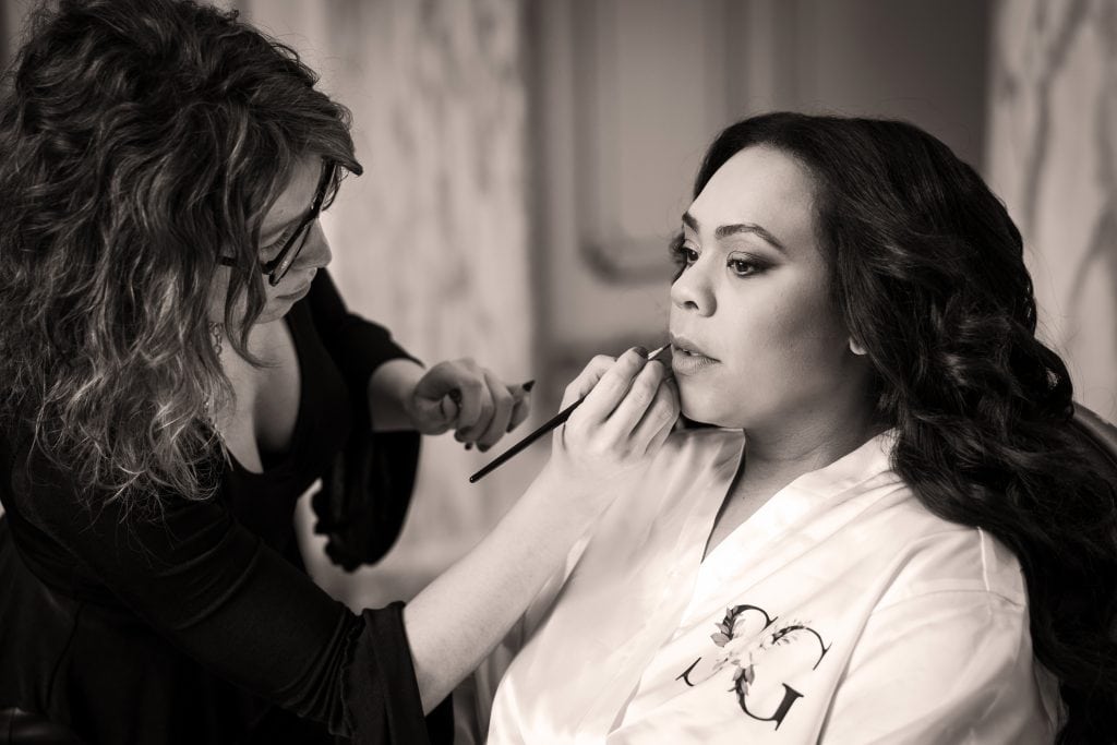 A makeup artist applies lipstick to a woman wearing a robe, who is seated and looking forward. The scene appears to be indoors, with a calm, focused atmosphere. Wedding Photo taken by Jennings Photography