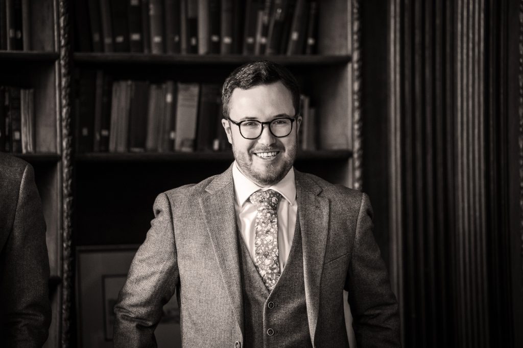 A man in a suit and tie stands smiling in front of a bookshelf in a formal indoor setting. Wedding Photo taken by Jennings Photography
