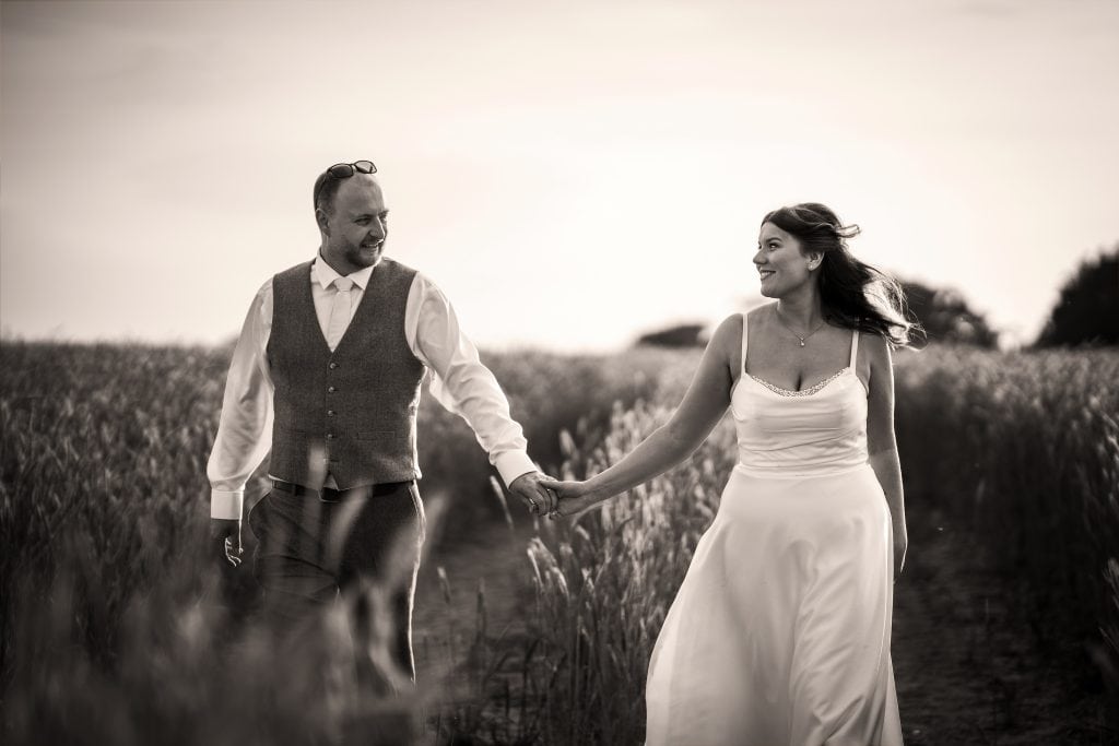 Ellie & James, dressed in formal attire, walk hand in hand through a field, smiling at each other. The image is captured in timeless black and white. Wedding Photo taken by Jennings Photography