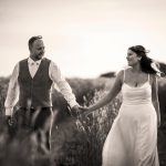 Ellie & James, dressed in formal attire, walk hand in hand through a field, smiling at each other. The image is captured in timeless black and white. Wedding Photo taken by Jennings Photography