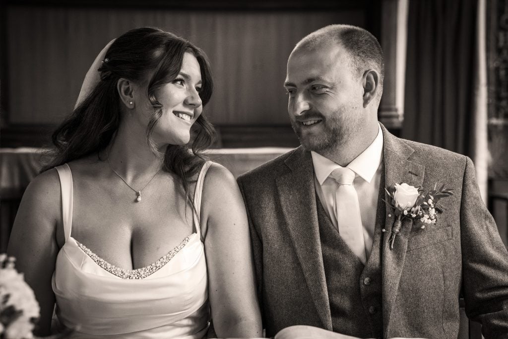 A bride and groom sit side by side indoors, dressed in wedding attire, smiling at each other. Wedding Photo taken by Jennings Photography