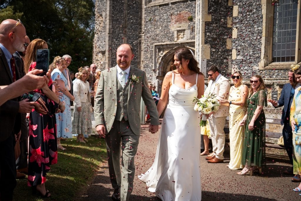 A bride and groom walk hand in hand outside a church as guests throw confetti and smile, celebrating their wedding on a sunny day. Wedding Photo taken by Jennings Photography