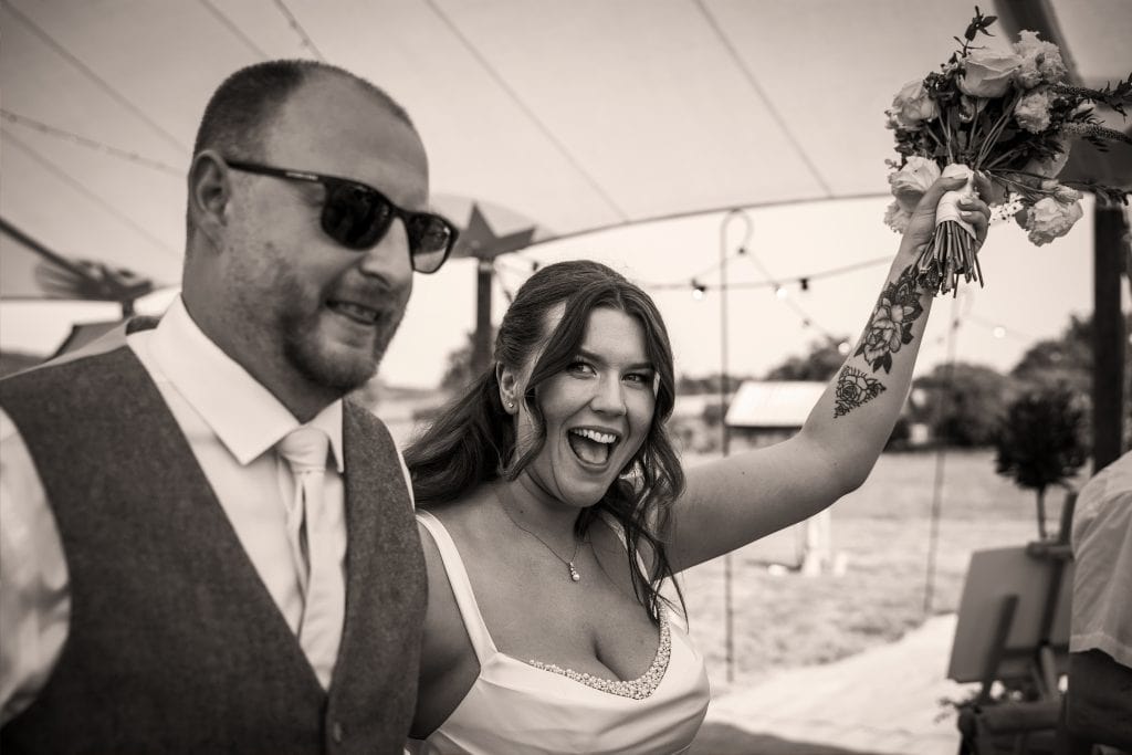 A smiling bride raises her bouquet while standing next to a man in sunglasses and a vest at an outdoor event, under a tent. Wedding Photo taken by Jennings Photography