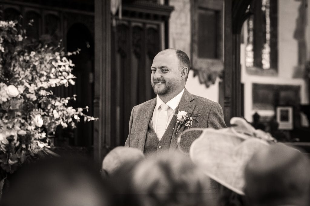 A man in a suit smiles while standing indoors at what appears to be a formal event, with blurred people and floral arrangements in the foreground. Wedding Photo taken by Jennings Photography