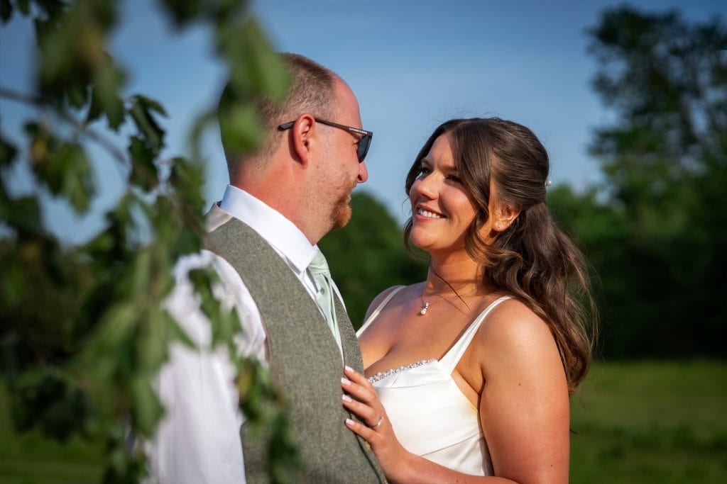 A couple stands outdoors, smiling at each other. The woman wears a white dress, and the man wears a vest and tie. Green foliage and a sunny sky are in the background. Wedding Photo taken by Jennings Photography