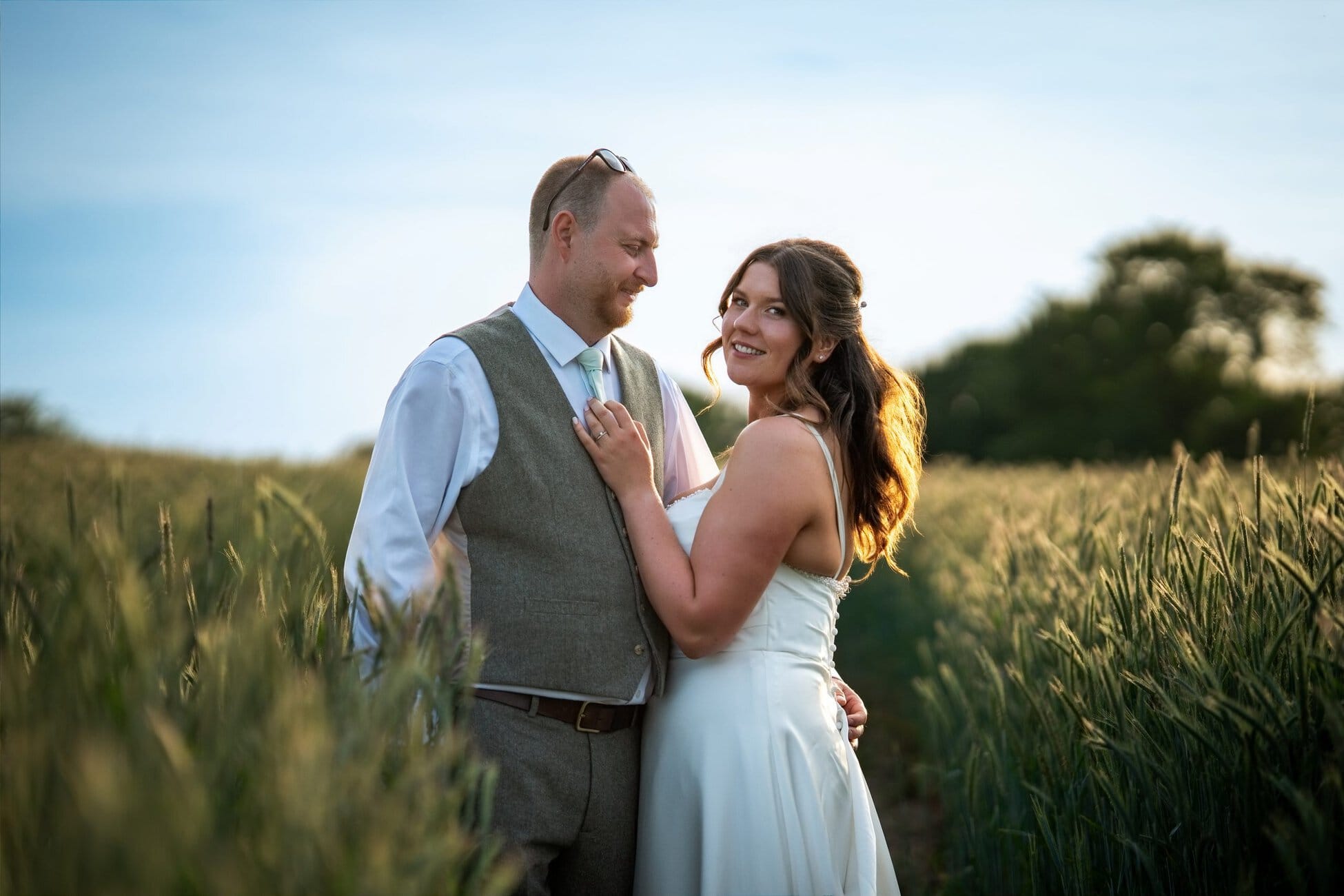 A couple stands close together in a field of tall grass, both dressed formally, with the woman smiling at the camera and the man looking at her. Trees and blue sky are visible in the background. Wedding Photo taken by Jennings Photography