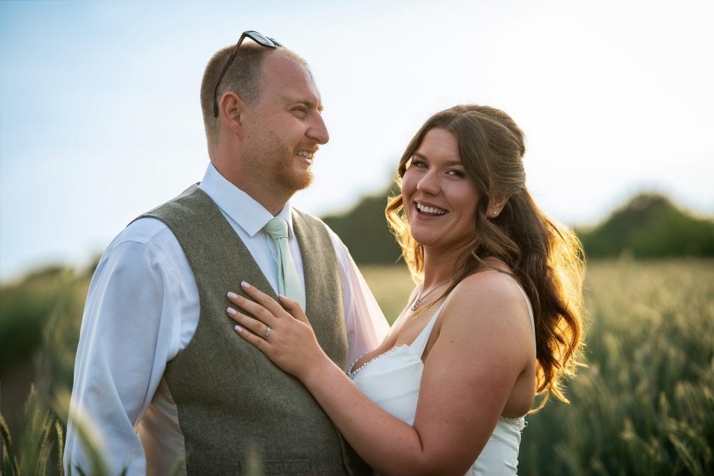 A couple stands in a field at sunset; the woman in a white dress smiles at the camera while the man looks at her, both appearing happy. Wedding Photo taken by Jennings Photography