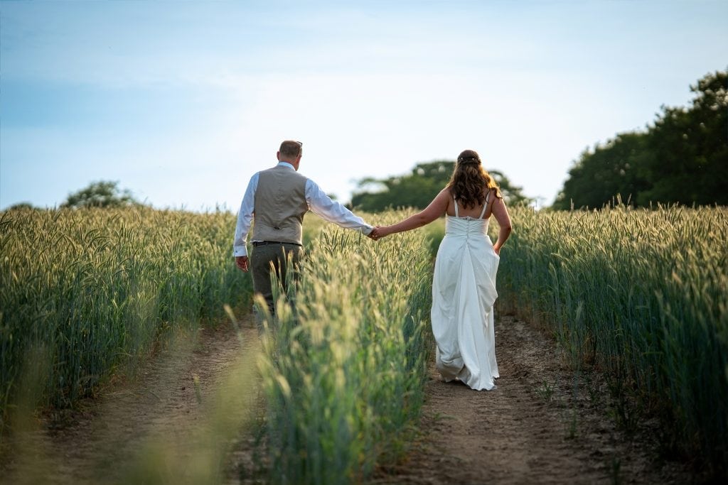 A bride and groom walk hand in hand through a grassy field under a blue sky, with trees in the background. Wedding Photo taken by Jennings Photography