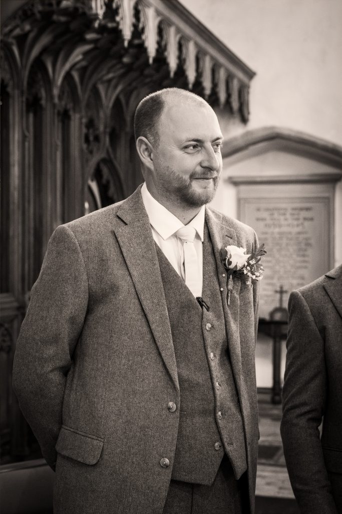A man in a suit stands indoors, likely at a formal event. He is smiling slightly, with hands behind his back. There is ornate woodwork and a plaque in the background. Black and white photo. Wedding Photo taken by Jennings Photography